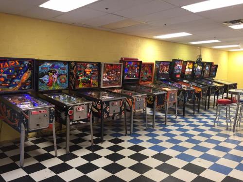A row of colorful pinball machines in a game room with a checkered floor and yellow walls.