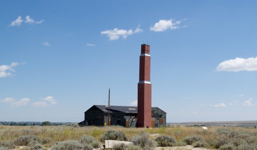 A tall red brick chimney stands beside an old, weathered building in a grassy landscape under a blue sky with clouds.