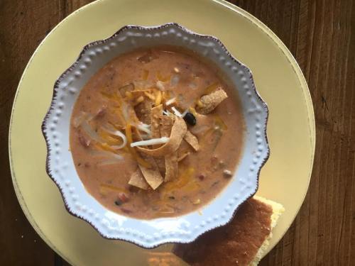A bowl of creamy soup topped with cheese, tortilla strips, and black beans, served with a piece of cornbread.
