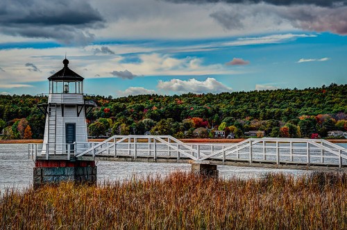 A lighthouse stands on a pier by a calm waterway, surrounded by autumn foliage and a cloudy sky.