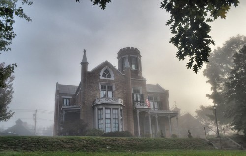 A grand, fog-covered mansion with a turret, surrounded by trees and a grassy area, evoking a mysterious atmosphere.