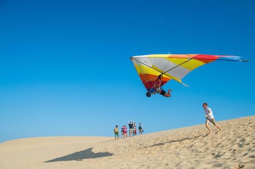 A person hang gliding over sand dunes, while a group of people cheer in the background under a clear blue sky.