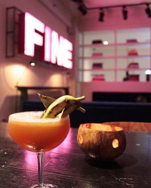 A cocktail with banana garnish sits on a table, with a wooden bowl and a glowing "FINE" sign in the background.