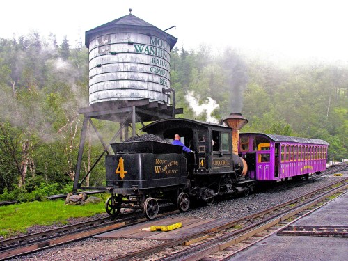 A vintage steam train with a purple passenger car stops near a water tower in a misty, green landscape.