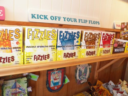 A shelf displaying colorful boxes of Fizzy instant sparkling drink tabs, with a sign above reading "Kick Off Your Flip Flops."