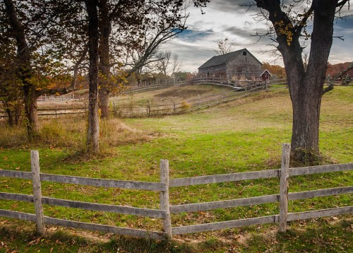 A rustic barn sits on a hillside, surrounded by trees and a wooden fence, under a cloudy sky.