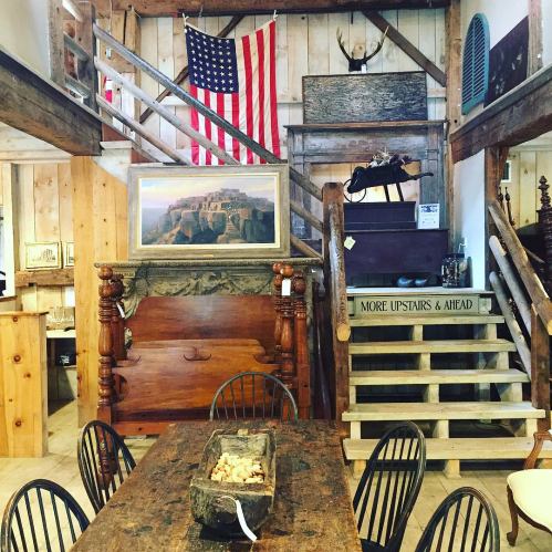 A rustic interior featuring wooden furniture, an American flag, and a staircase leading to an upper level.