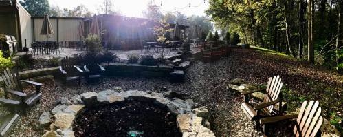 A serene outdoor space with wooden chairs, a fire pit, and a patio surrounded by trees and sunlight.