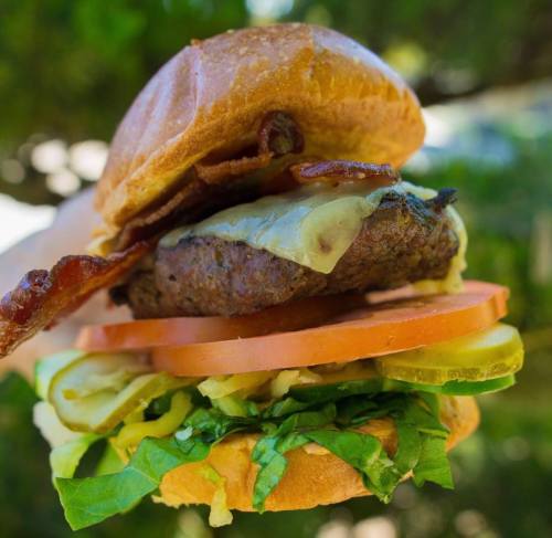 A close-up of a juicy burger with bacon, cheese, lettuce, tomato, and pickles, held against a blurred green background.