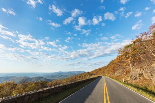 A winding road through autumn-colored mountains under a blue sky with scattered clouds.