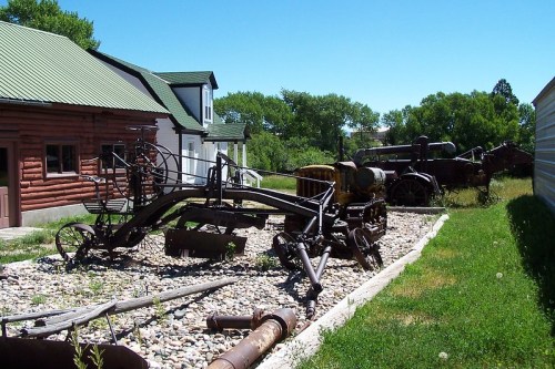 Rusty vintage farm equipment displayed outdoors near a log cabin and greenery under a clear blue sky.