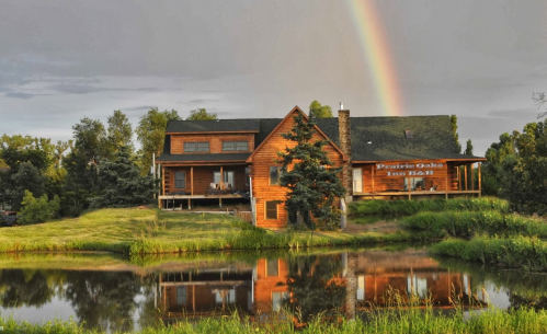 A wooden house by a pond, reflecting in the water, with a rainbow in the sky above. Lush greenery surrounds the scene.