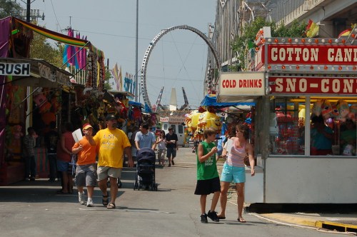 A lively fair scene with colorful stalls, people walking, and a Ferris wheel in the background. Cotton candy and drinks are featured.