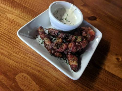 A plate of bacon-wrapped food with a small bowl of dipping sauce and a garnish of herbs on a wooden table.