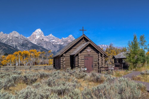 A rustic log cabin church surrounded by autumn trees and mountains under a clear blue sky.