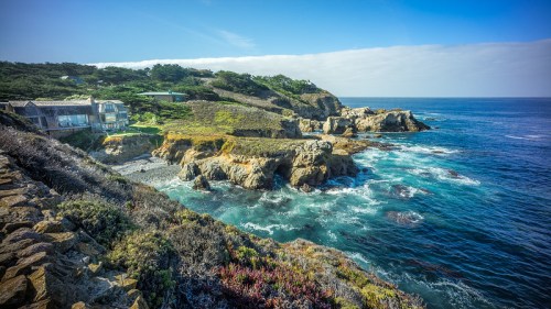 Coastal view featuring rocky cliffs, waves crashing, and a house nestled among greenery under a clear blue sky.