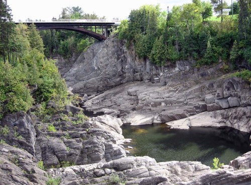 A rocky landscape with a river pool, surrounded by trees and a bridge overhead.