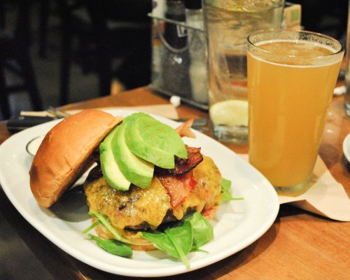 A juicy burger topped with avocado, bacon, and greens, served with a glass of light beer on a wooden table.