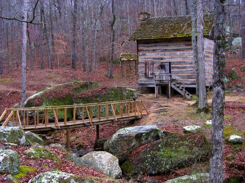 A rustic wooden cabin beside a small stream, with a bridge and surrounded by trees in a forested area.