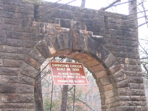 A stone archway with a sign reading "Swinging Bridge Built in 1939" and safety instructions about bridge use.