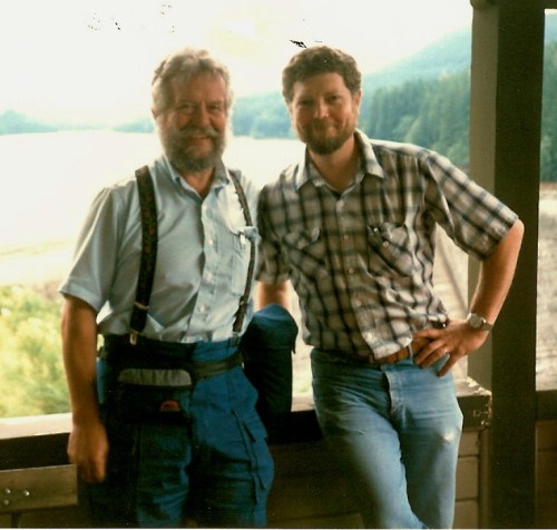 Two men stand together on a balcony overlooking a scenic landscape, smiling at the camera.