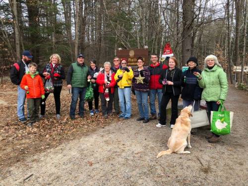 A group of people in festive attire poses together at a trailhead, with a dog and holiday decorations nearby.