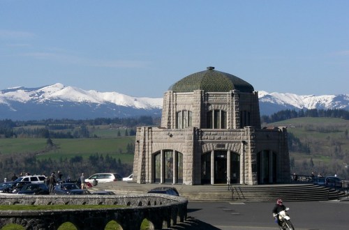 A stone building with a dome, surrounded by green hills and snow-capped mountains under a clear blue sky.