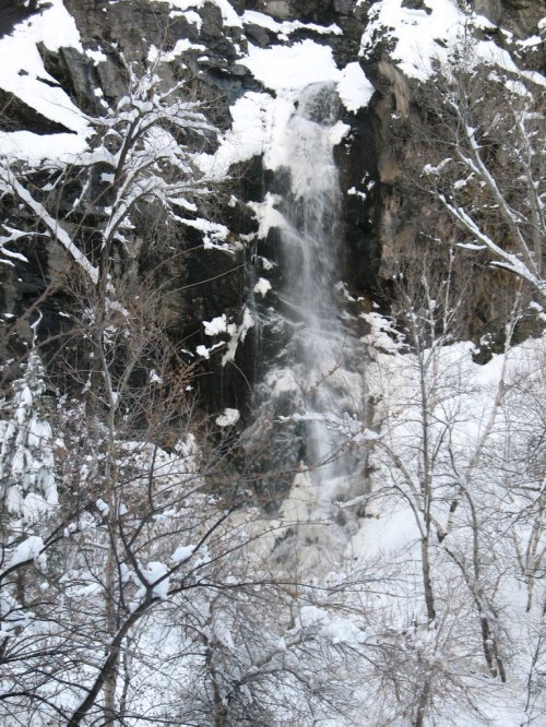 A snowy landscape featuring a waterfall cascading down rocky cliffs, surrounded by bare trees.