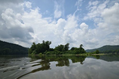 A serene lake scene with lush greenery, an island, and clouds reflecting on the water's surface.
