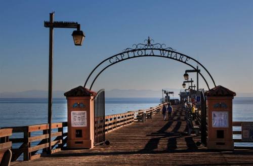 A wooden pier with an archway reading "Capitola Wharf," leading to calm waters and distant mountains under a clear sky.