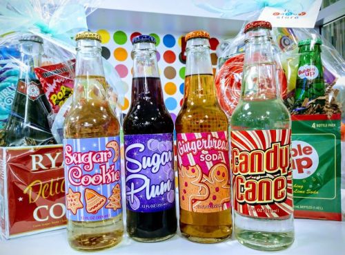 A colorful display of festive soda bottles: Sugar Cookie, Sugar Plum, Gingerbread, and Candy Cane, with gift baskets in the background.