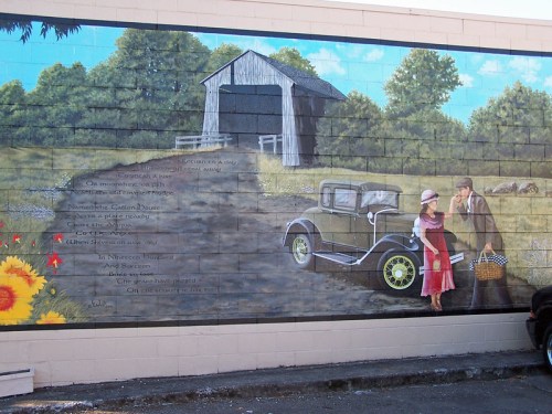 A mural depicting a vintage car, a couple, and a scenic rural landscape with flowers and a barn.