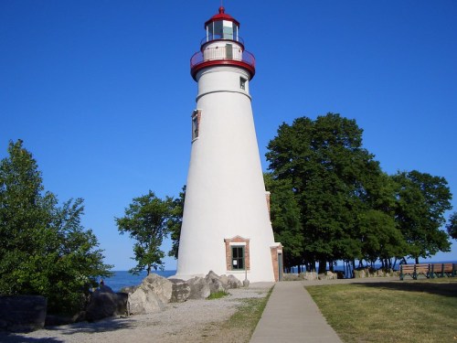 A tall white lighthouse with a red top, surrounded by trees and a clear blue sky, near a pathway and rocky shoreline.