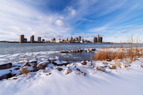 A snowy landscape with rocks in the foreground and a city skyline under a blue sky in the background.