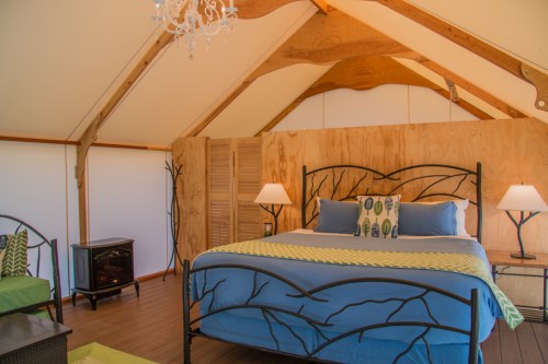 Cozy glamping tent interior with a bed, lamps, and decorative accents, featuring wooden walls and a chandelier.