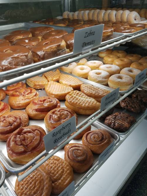 A display case filled with various donuts, including glazed, filled, and specialty varieties like Zebra and Raspberry Rosebuds.
