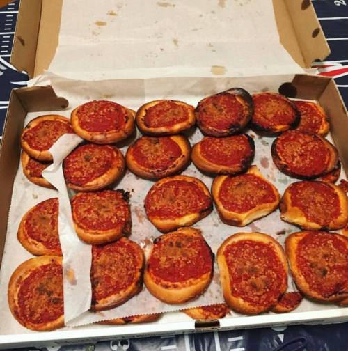 A pizza box filled with small, round pizzas, some burnt, topped with tomato sauce and herbs.