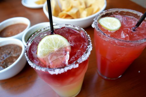 Two colorful cocktails with lime slices and salt rims, accompanied by bowls of salsa and tortilla chips in the background.