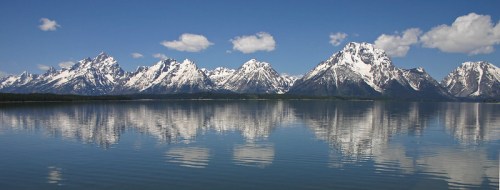 A panoramic view of snow-capped mountains reflecting in a calm lake under a clear blue sky with fluffy clouds.