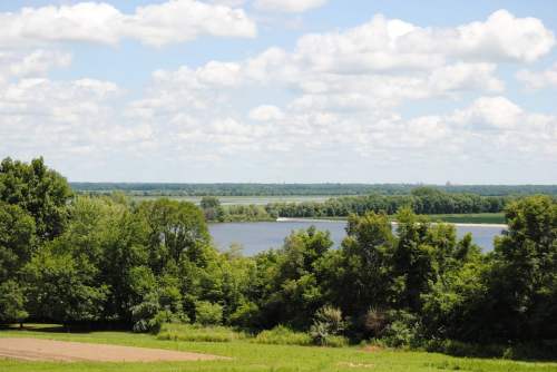 A scenic view of a river surrounded by lush greenery and fluffy clouds in a blue sky.