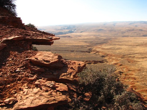 A rocky ledge overlooks a vast, arid landscape with rolling hills and sparse vegetation under a clear blue sky.