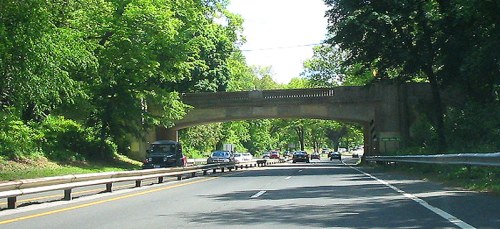 A tree-lined road with a stone bridge overhead, featuring vehicles traveling in both directions.