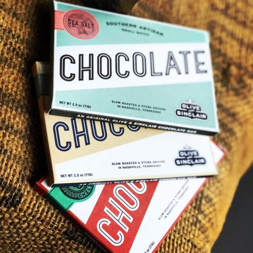 Three chocolate bars with colorful packaging, stacked on a textured surface. Labels include "Sea Salt" and "Southern Artisan."