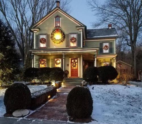 A charming two-story house decorated with wreaths and lights, surrounded by snow and neatly trimmed bushes.
