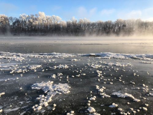 A frozen lake with mist rising, surrounded by trees frosted with snow under a clear blue sky.