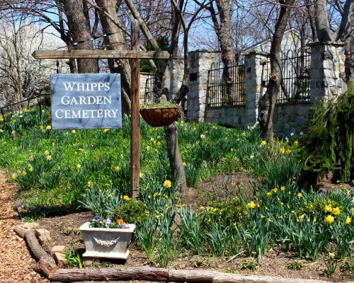 Sign for Whipps Garden Cemetery surrounded by blooming flowers and trees in a serene outdoor setting.