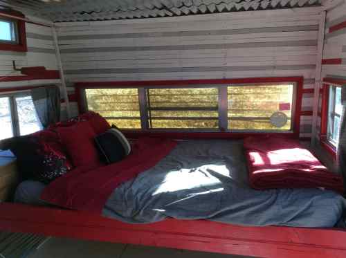Cozy interior of a tiny home with a bed dressed in red and gray linens, large windows, and wooden accents.