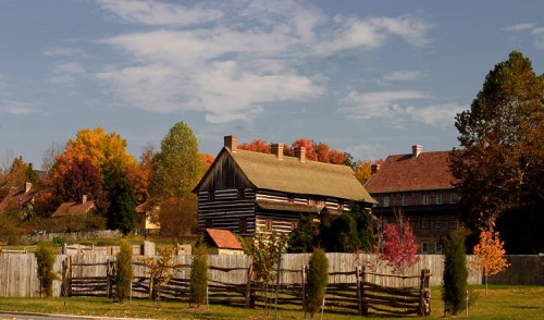 A rustic log cabin surrounded by colorful autumn trees and a wooden fence under a blue sky with scattered clouds.