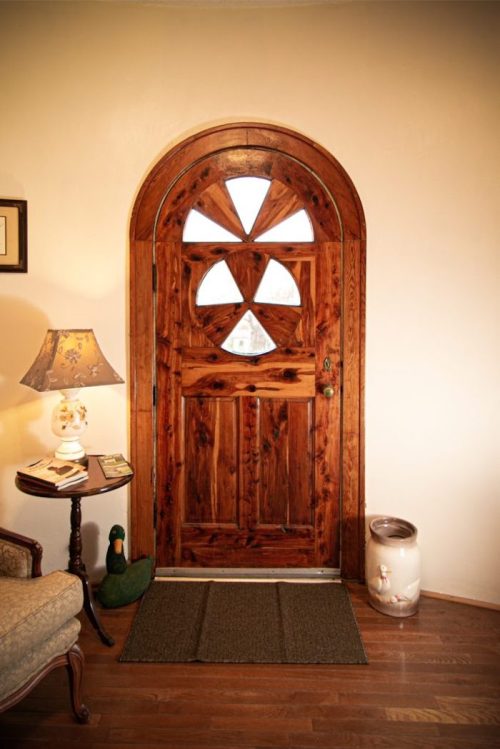 A wooden arched door with a decorative window, next to a lamp and a small table, on a hardwood floor.