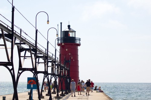 A red lighthouse on a pier with people walking along it, overlooking a calm sea under a bright sky.
